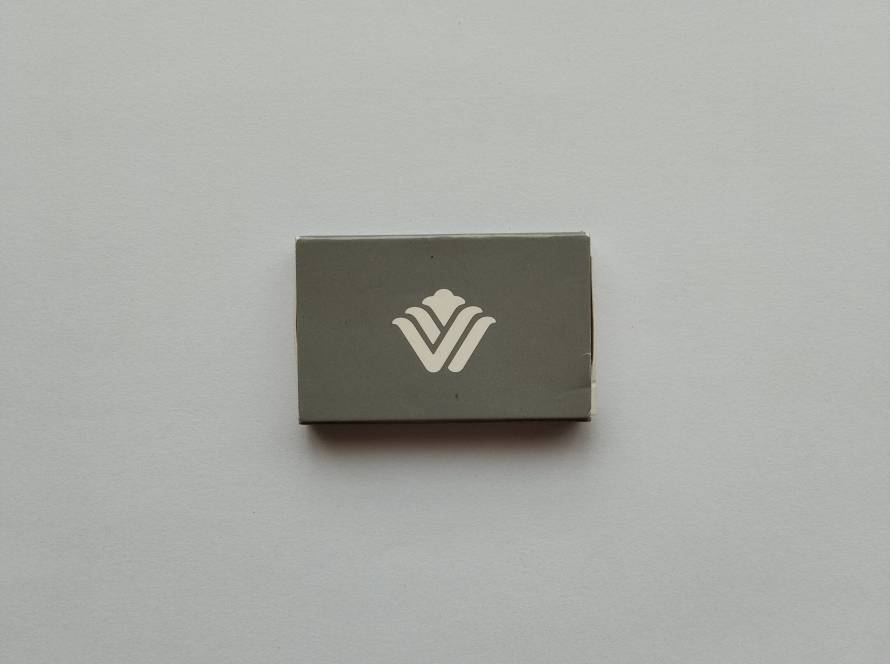 Grey minimalist hotel matchbox featuring white stylized “V” logo on both sides. Clean modern branding design.