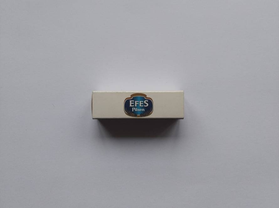 “Efes Pilsen” side-logo promotional matchbox; cream background with centered classic blue-gold Efes emblem. Clean minimalist design.
