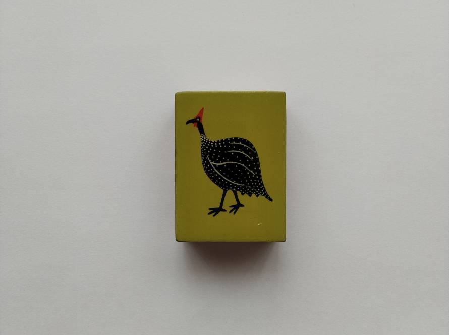 Minimalist yellow matchbox case featuring a stylized guinea fowl with dotted plumage and red crest. Bold flat-color design with modern graphic simplicity.