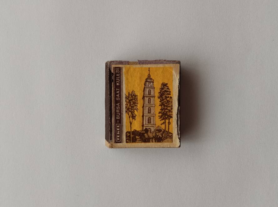 Tekel “Bursa Saat Kulesi” illustrated matchbox. Yellow-toned architectural drawing with vertical TEKEL band on the side. Turkish landmark series collectible.