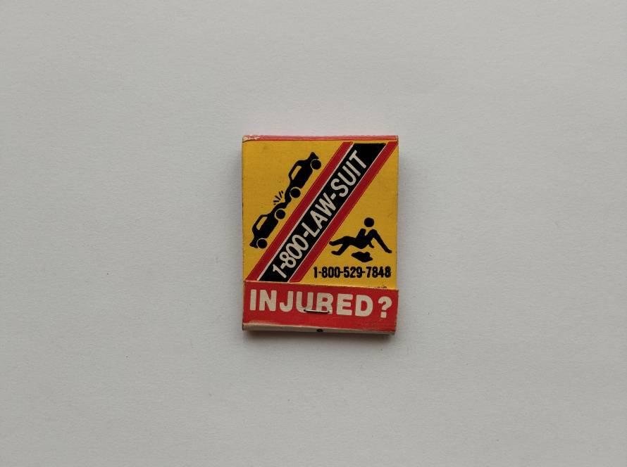 Bold accident and injury legal advertisement matchbox with hotline number and caution-style graphics, typical of U.S. promotional items.