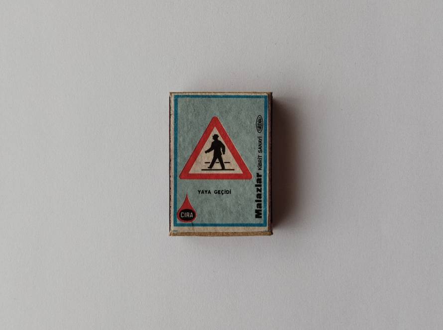 Vintage ÇIRA Malazlar Kibrit Sanayi matchbox featuring “Yaya Geçidi” (pedestrian crossing) warning road sign design with turquoise background and red triangle border.