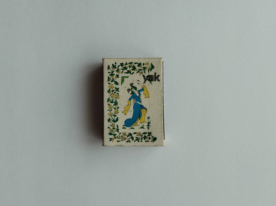 Vintage “Yak” branded matchbox featuring dancing woman illustration surrounded by floral decorative border.