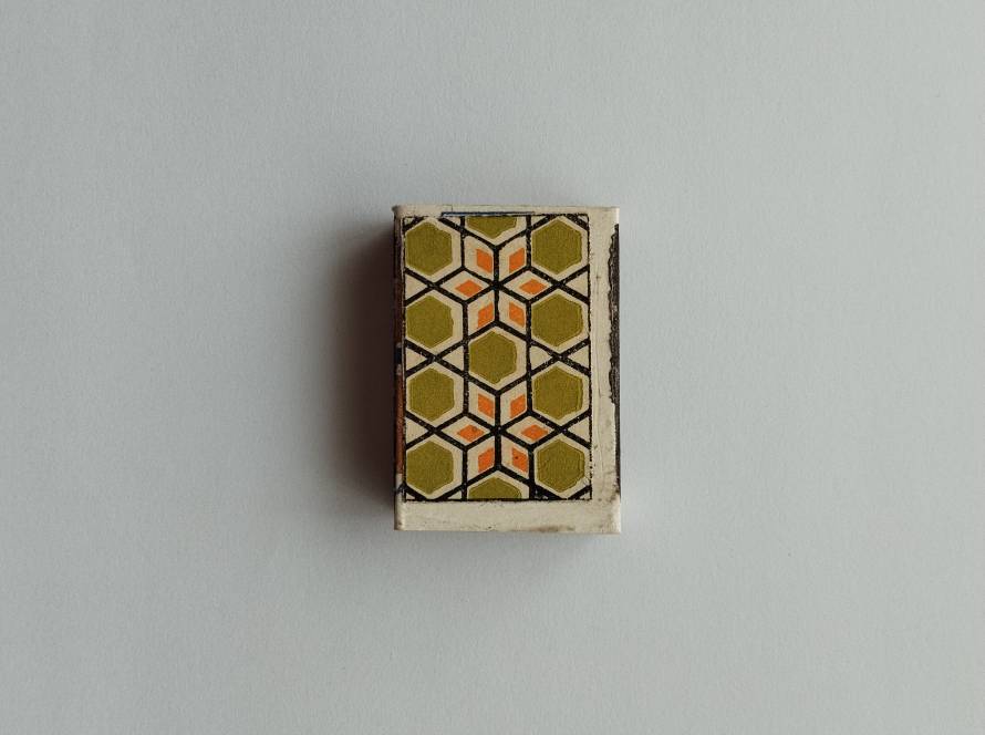 Abstract geometric honeycomb-style pattern matchbox in olive green, orange, and black tones.