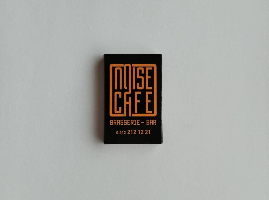 Promotional matchbox for Noise Cafe Brasserie & Bar, featuring modern black and orange branding with contact information.